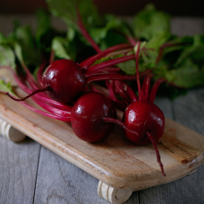 Close up of Gemini beet roots and tops showing dark red smooth globe shaped roots and bright green leaves with magenta veins on a wooden board