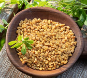 Top-down view of fenugreek seeds in a wooden bowl with a small green sprig on weathered wood background