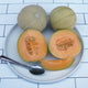 Close-up of Minnesota Midget cantaloupes, two whole and one halved, showcasing deep orange flesh and netted rind on a ruler-marked background