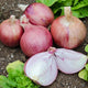 A group of red blush onions with one cut in half, showing the interior. The onions are on a bed of soil with green leaves around.