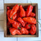 A wooden crate filled with bright red NOTTA HOTTA peppers with a waxy appearance, displayed on a white tiled background.