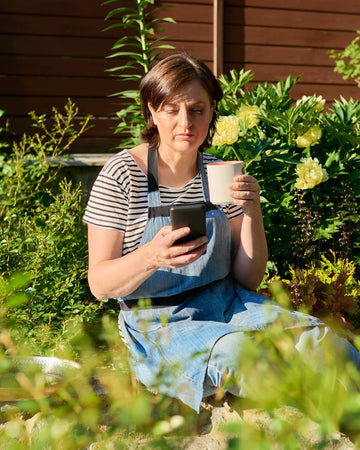 Woman in a garden holding a phone and a mug, surrounded by greenery.