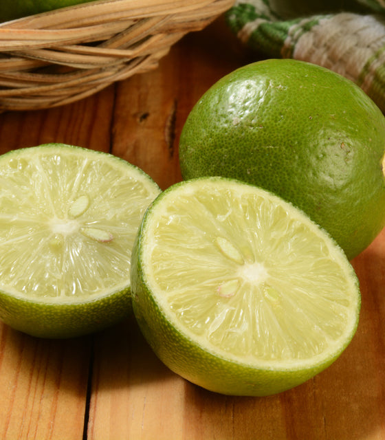 Close-up of Thornless Mexican Key lime showing two cut halves and one whole fruit with glossy thin skin on a wooden surface