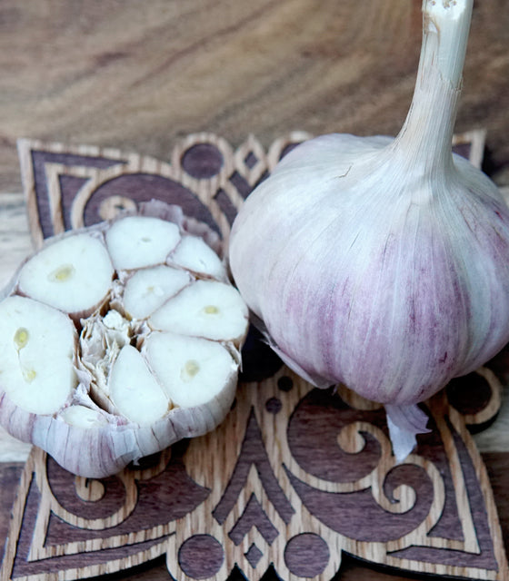 A whole Krandasger Red garlic bulb and a halved garlic bulb placed on a decorative wooden board over a wood surface.