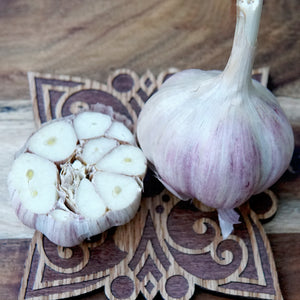 A whole Krandasger Red garlic bulb and a halved garlic bulb placed on a decorative wooden board over a wood surface.