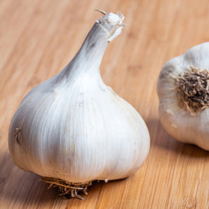 A close-up of a whole blue music garlic bulb on a light wooden surface, with another garlic bulb partially visible in the background.