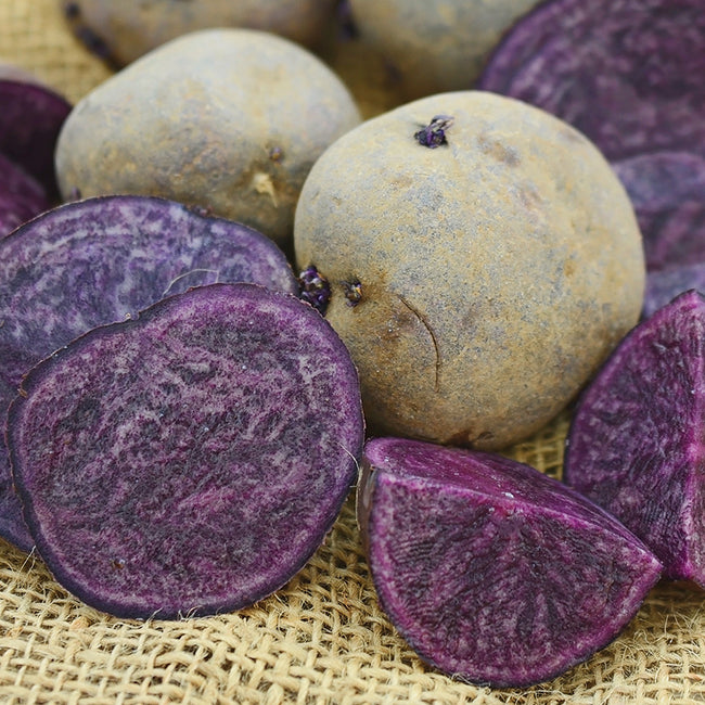 Whole and sliced purple majesty potatoes on a burlap sack surface