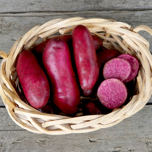 Overhead view of Raspberry seed potatoes in a woven basket showing deep red skin and a sliced tuber with bright red flesh on wooden boards