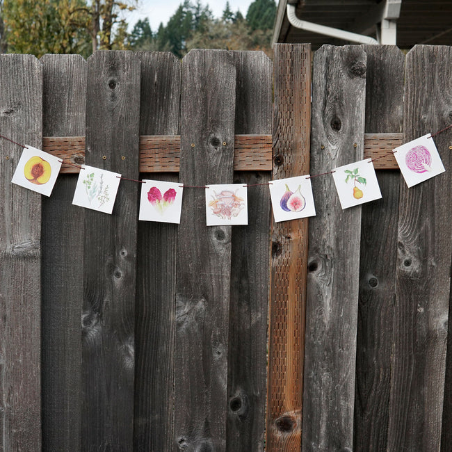 Front view of seven watercolor produce canvas flags hand strung on hemp cord hanging on a wooden fence