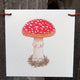 Front view of mushroom flag showcasing a red-spotted watercolor mushroom on white canvas hung with clips against a wooden background
