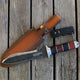 Angled view of The Hero Hori tool with 7.5-inch double-sided blade, cocobolo handle, and leather sheath on wood
