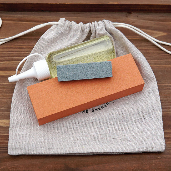 Top-down view of tool sharpening kit showing two sharpening stones (orange fine, gray coarse), a small oil bottle on a linen drawstring bag