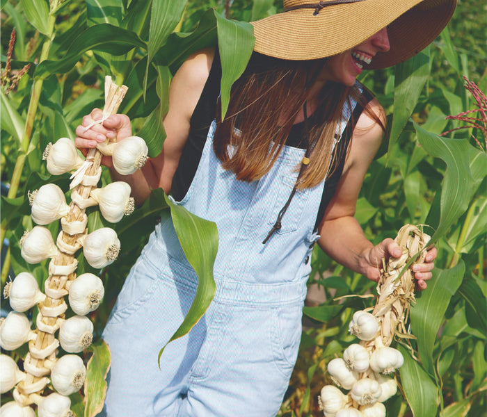 Woman in a straw hat and denim overalls holding two braided garlic bunches in a garden, smiling