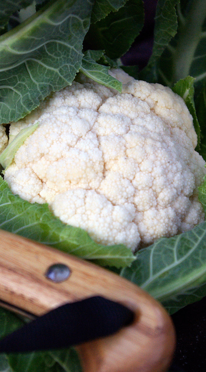 Close-up of a cauliflower head with green leaves and a knife in the foreground.