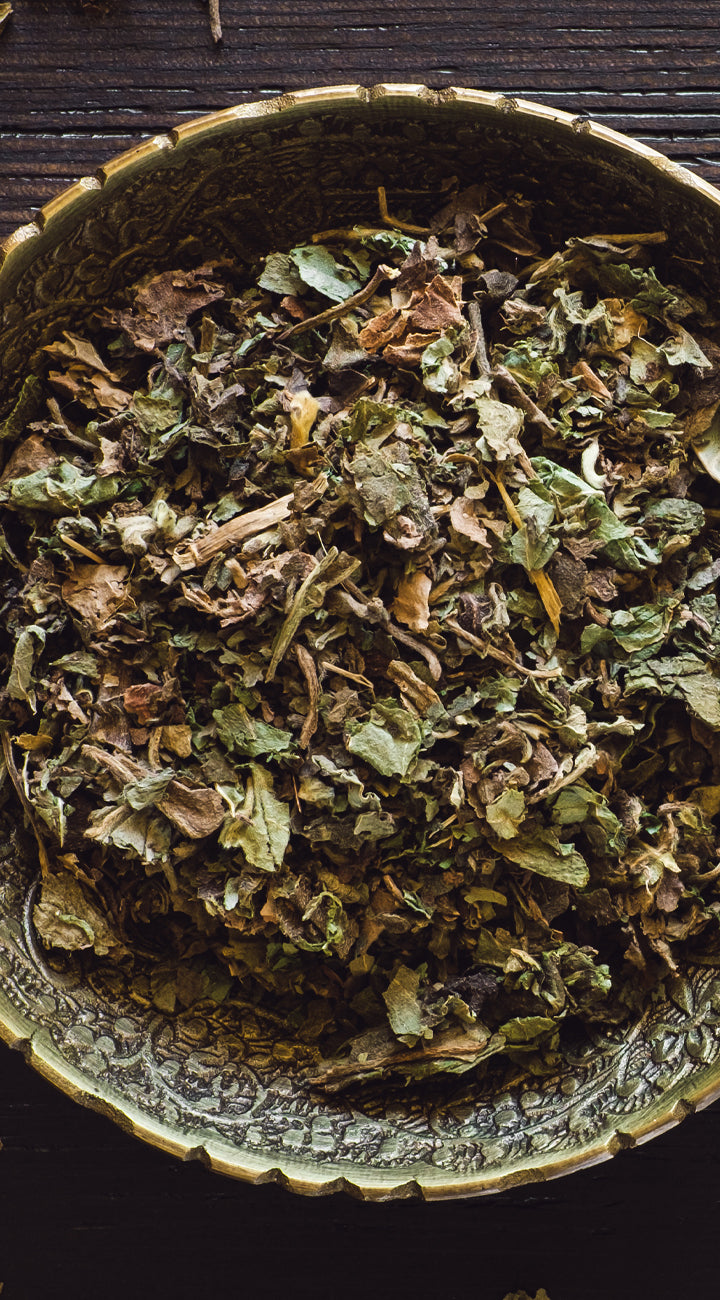 Dry green patchouli leaves in a decorative metal bowl on a dark wooden surface