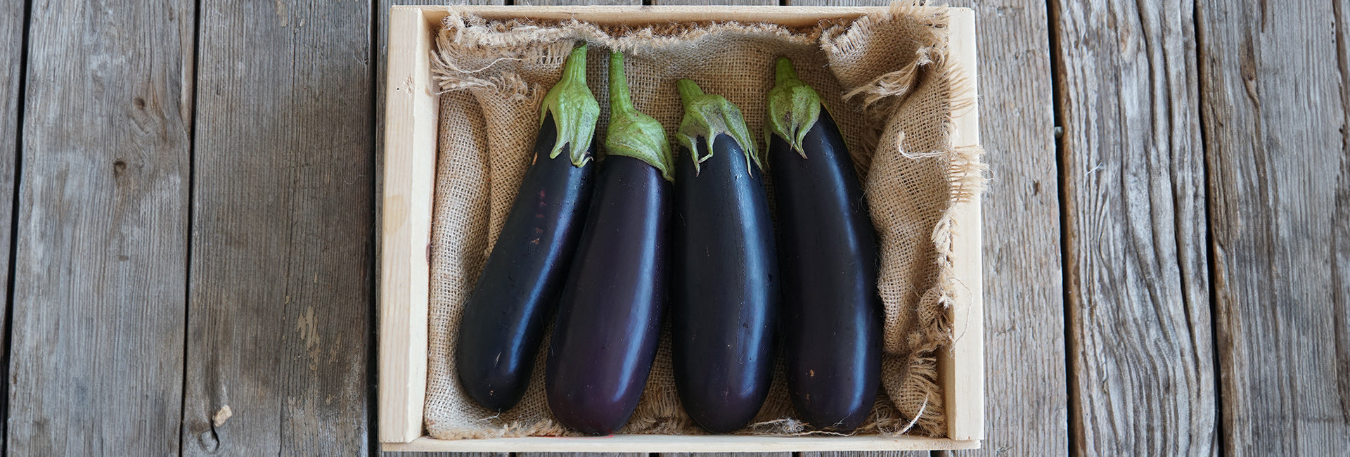 Top-down view of four glossy deep-purple eggplants with green calyxes in a wooden crate lined with burlap on weathered wood