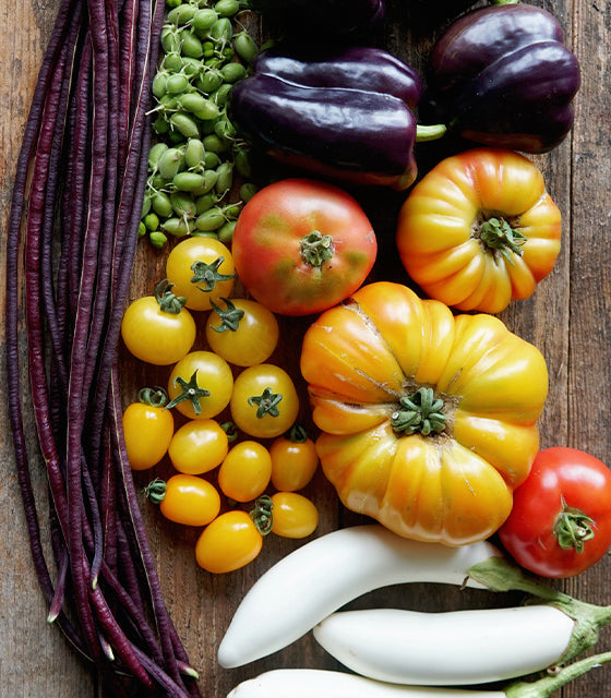 A flat-lay arrangement of fresh, colorful vegetables on a rustic wooden surface, including dark purple bell peppers, long purple beans, green beans, yellow and red tomatoes, and white eggplants.