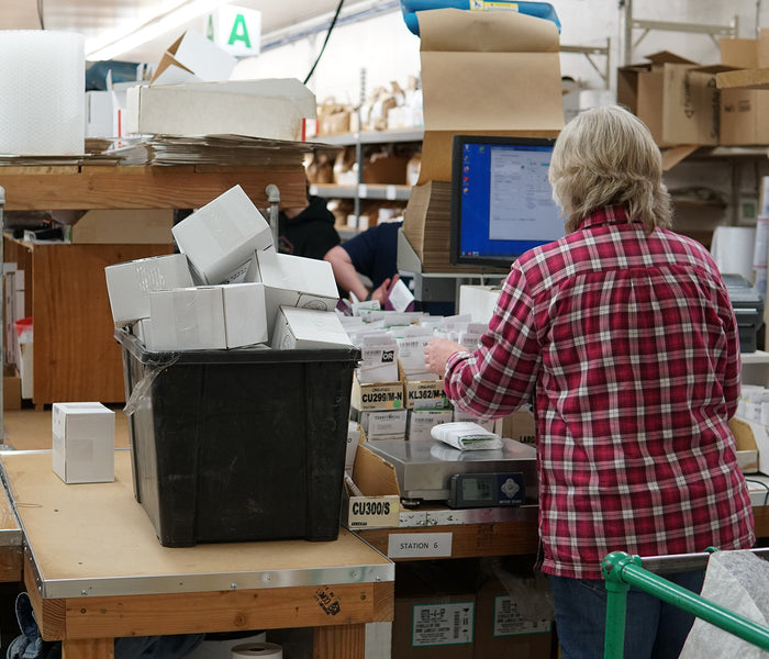 Person working at a packaging station with boxes and a computer in a warehouse setting.