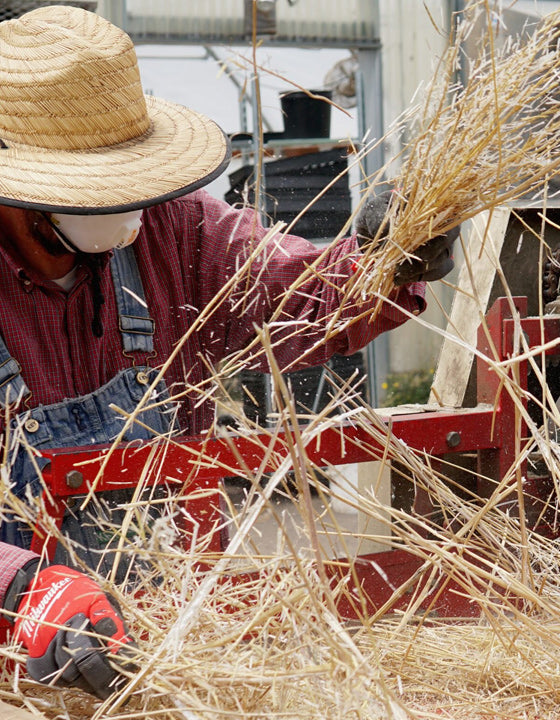 A person wearing a straw hat, red plaid shirt, blue denim overalls, and red gloves handles a bundle of dry straw in front of a red machine.