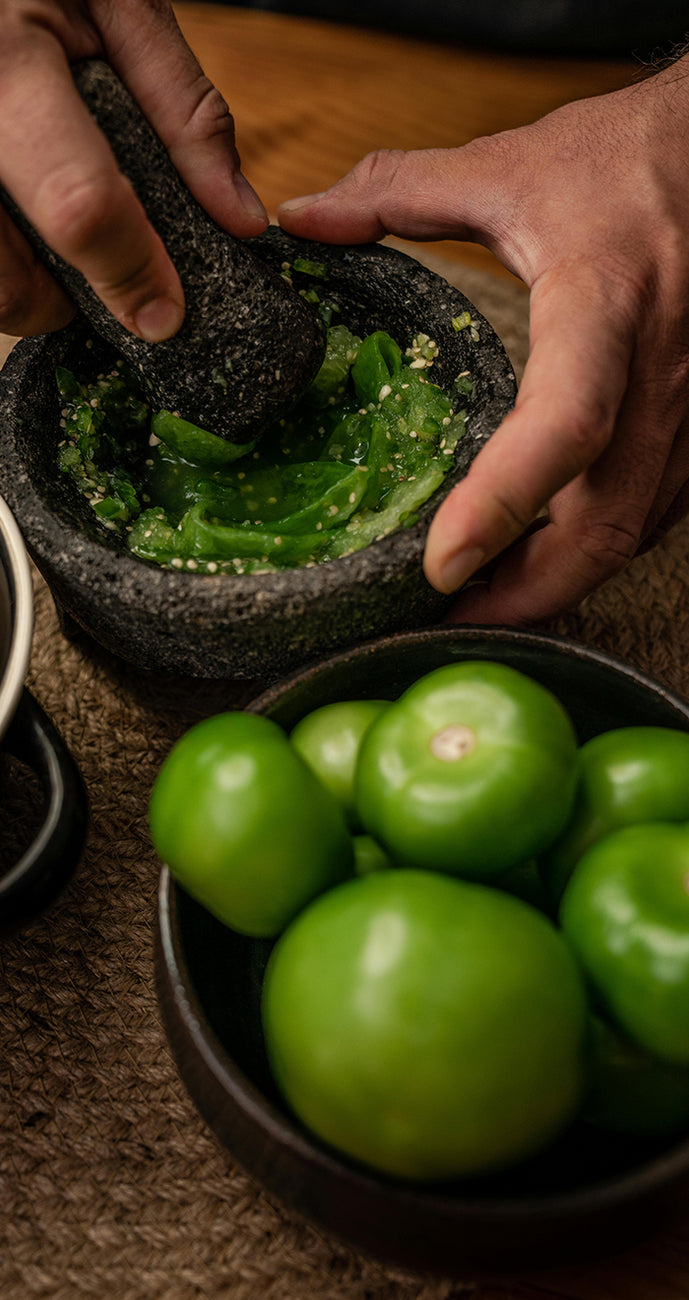 Person using a mortar and pestle with green tomatillos on a wooden surface