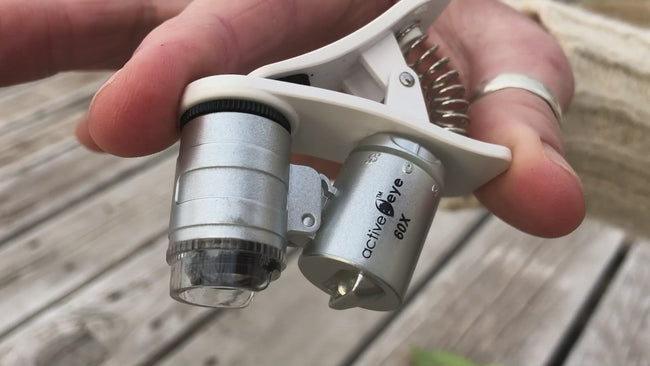 Close-up of Universal Mobile Phone Microscope 60X clipped in a hand showing the 60x lens and built-in LED against a wooden deck