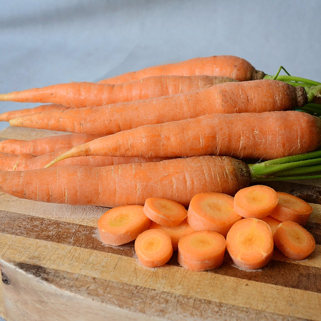 Close up of Bolero carrots, bright orange whole carrots with green tops and sliced rounds on a wooden cutting board