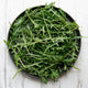 Overhead view of Bellezia arugula showcasing dark green, deeply serrated leaves on a round black plate over white wood