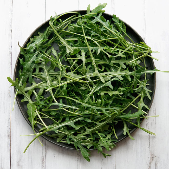 Overhead view of Bellezia arugula showcasing dark green, deeply serrated leaves on a round black plate over white wood