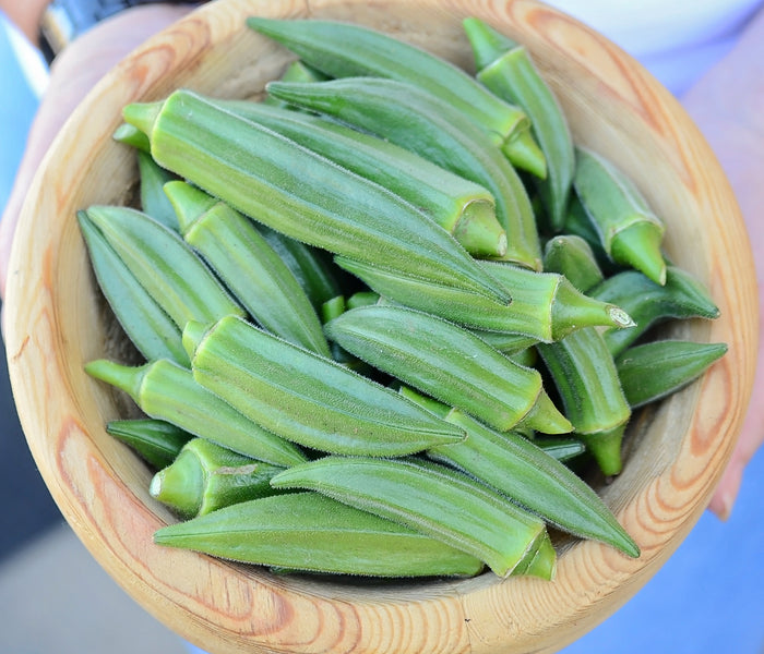 Top-down view of Jambalaya okra pods in a wooden bowl showcasing 5-ridged green, meaty pods held by a person