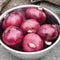 Top-down view of Monastrell red onions in a stainless steel bowl showing lustrous dark red, slightly flattened bulbs on a stone surface