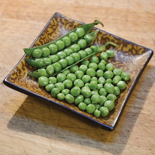 Angled top view of Canoe pea pods and shelled bright green peas on a patterned square dish set on a wooden cutting board