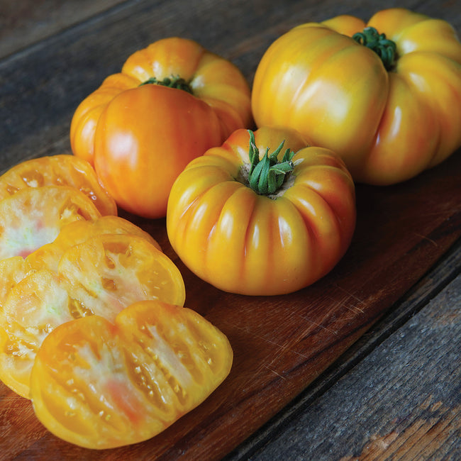Close-up of three pleated orange-yellow Buffalosun tomatoes, one sliced to show orange flesh on a wooden board