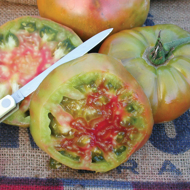 Close-up of Ananas Noire tomato halves showcasing green-and-red marbled flesh and glossy ribbed skin on burlap with small knife