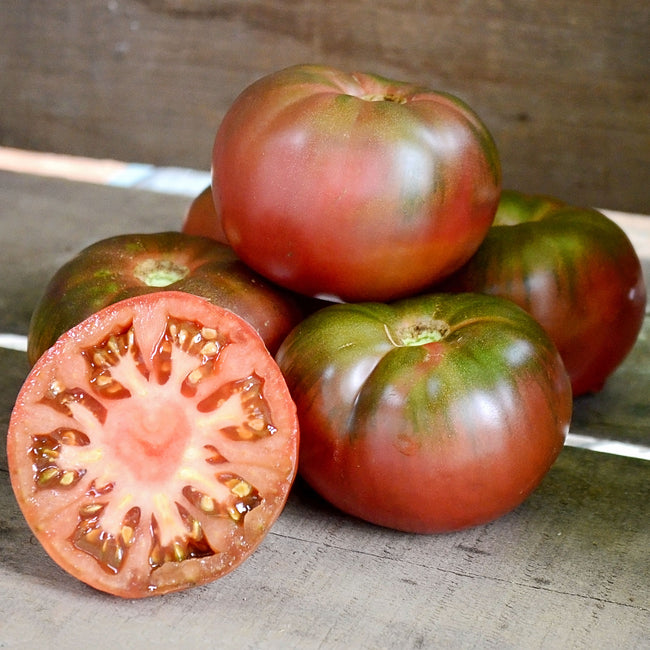 Front view of Cherokee Purple tomatoes showing flattened purple-red fruits with one sliced to reveal pink marbled interior on white background