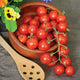 Close-up of Lizzano cherry tomatoes on the vine showing bright red 1-inch fruits with a wooden bowl, wooden spoon, and gray cloth background