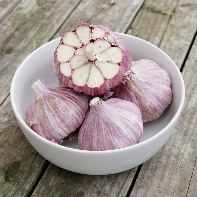 Overhead view of Chesnok Red garlic heads, one halved to show creamy cloves and purple-striped wrappers in a white bowl on weathered wood