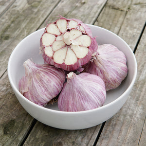 Overhead view of Chesnok Red garlic heads, one halved to show creamy cloves and purple-striped wrappers in a white bowl on weathered wood