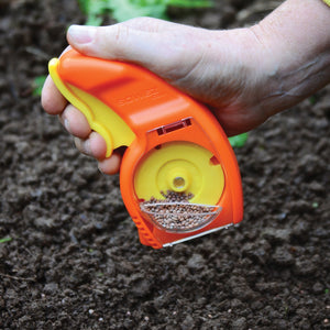 Handheld Sowez seeder showing clear hopper window, orange body, and seed-dispensing disk over soil