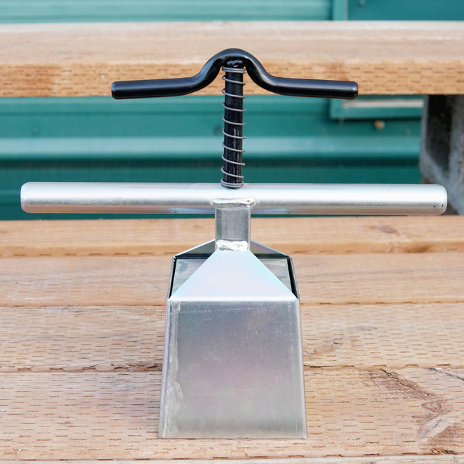 Angled front view of 4-inch soil blocker tool showcasing square metal mold and T-handle clamped to a wooden bench