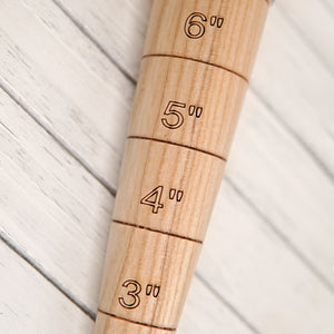 Angled side view of Dewit ash-wood dibber showcasing sharp brass tip and engraved inch markings on a light background