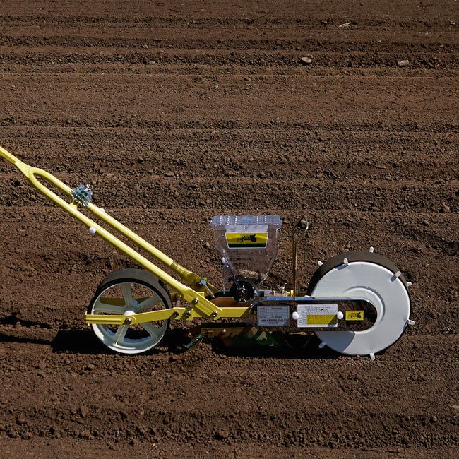 Side view of Jang one-row push seeder showing clear seed hopper, adjustable yellow frame, and press wheel on tilled soil